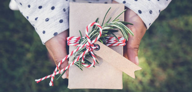 Someone holding a rectangle present wrapped in brown paper and decorated with string and foliage 