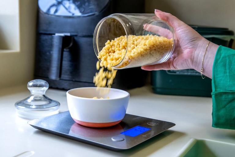 Hand pouring pasta into a bowl on kitchen scales.