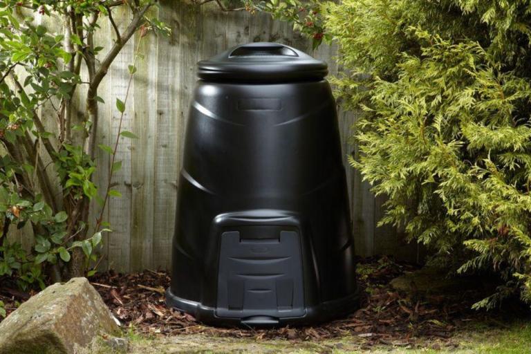 Black compost bin against a fence in a garden.