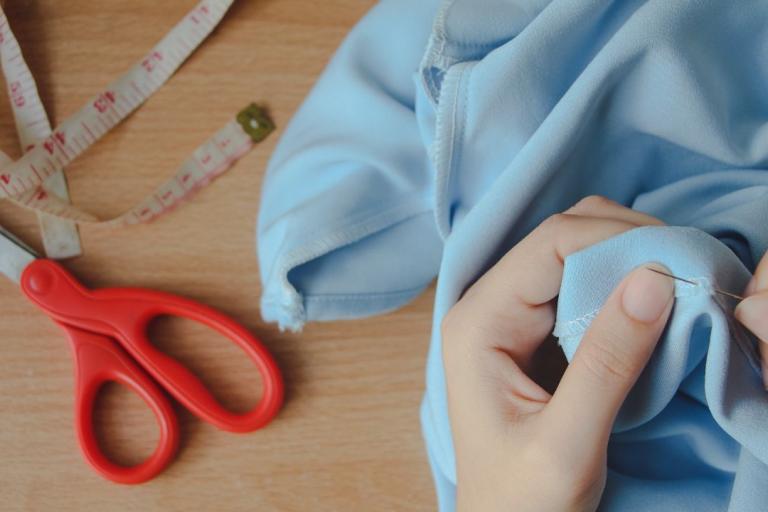 A pair of hands sewing some blue fabric with scissors and a measuring tape on the table.