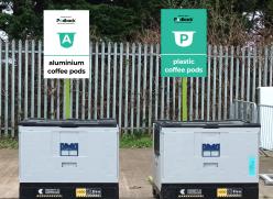 Two large, grey containers each with a rectangular sign above them. The white sign on the left says, “Recycle with Podback, The Pod Recycling Service: Aluminium Coffee Pods”. The turquoise sign on the right says, “Recycle with Podback, The Pod Recycling Service: Plastic Coffee Pods”. 