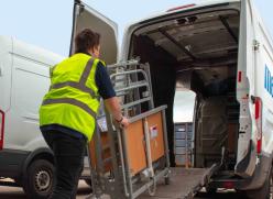 A man wearing a yellow hi-vis vest loading a bed frame onto a van, via a ramp.