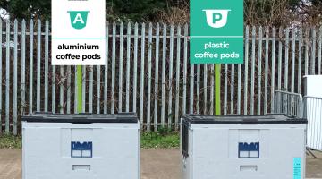 Two large, grey containers each with a rectangular sign above them. The white sign on the left says, “Recycle with Podback, The Pod Recycling Service: Aluminium Coffee Pods”. The turquoise sign on the right says, “Recycle with Podback, The Pod Recycling Service: Plastic Coffee Pods”. 