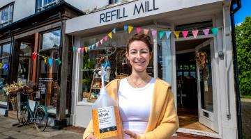 Vicki Daws, founder of Refill Mill, standing outside the shop holding a large soap dispenser. 